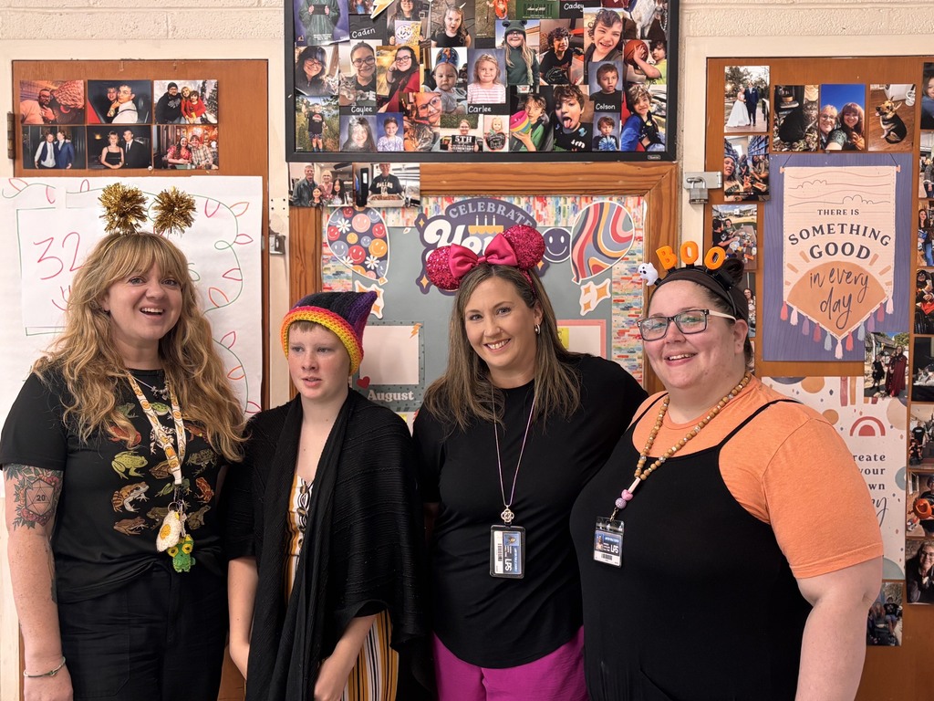 Four women pose in front of a bulletin board with photos and text. Each wears a hat or headpiece.