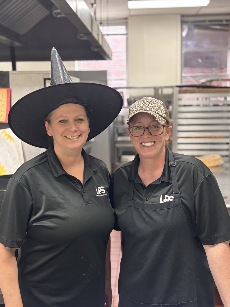 Two women wearing black aprons and polo shirts, one with a black witch hat, smiling for the camera in a kitchen.