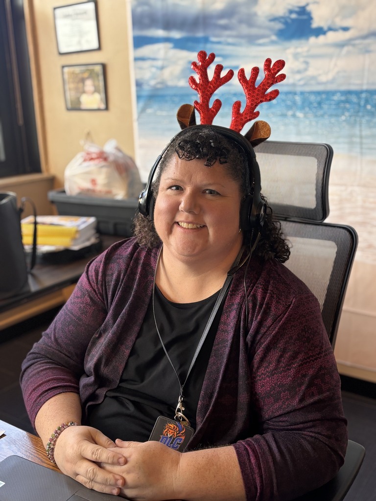 Woman sitting at desk with reindeer headband, headphones, and ID badge, smiling at the camera.