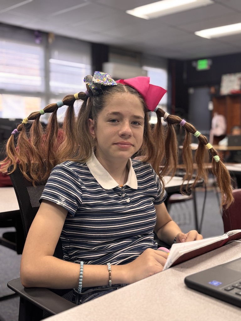 Girl seated in classroom, wearing pigtails tied up, reading a book and using a laptop.