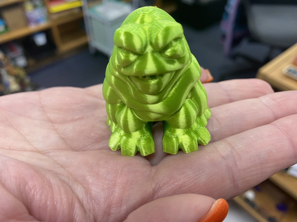 Hand holding a green toy figure with a large, round head, tiny limbs, and a large mouth.