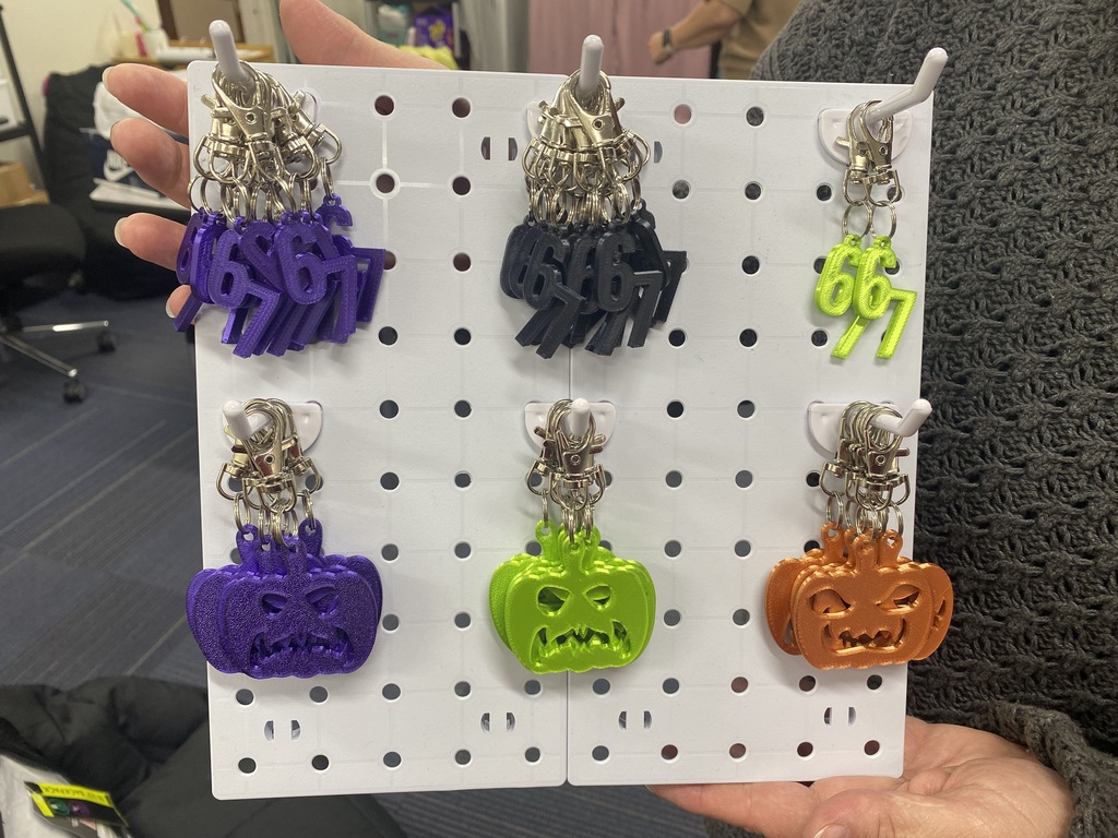 A person holds a white board with a grid and colorful Halloween-themed keychains attached. The keychains have Halloween pumpkins and bats on them.