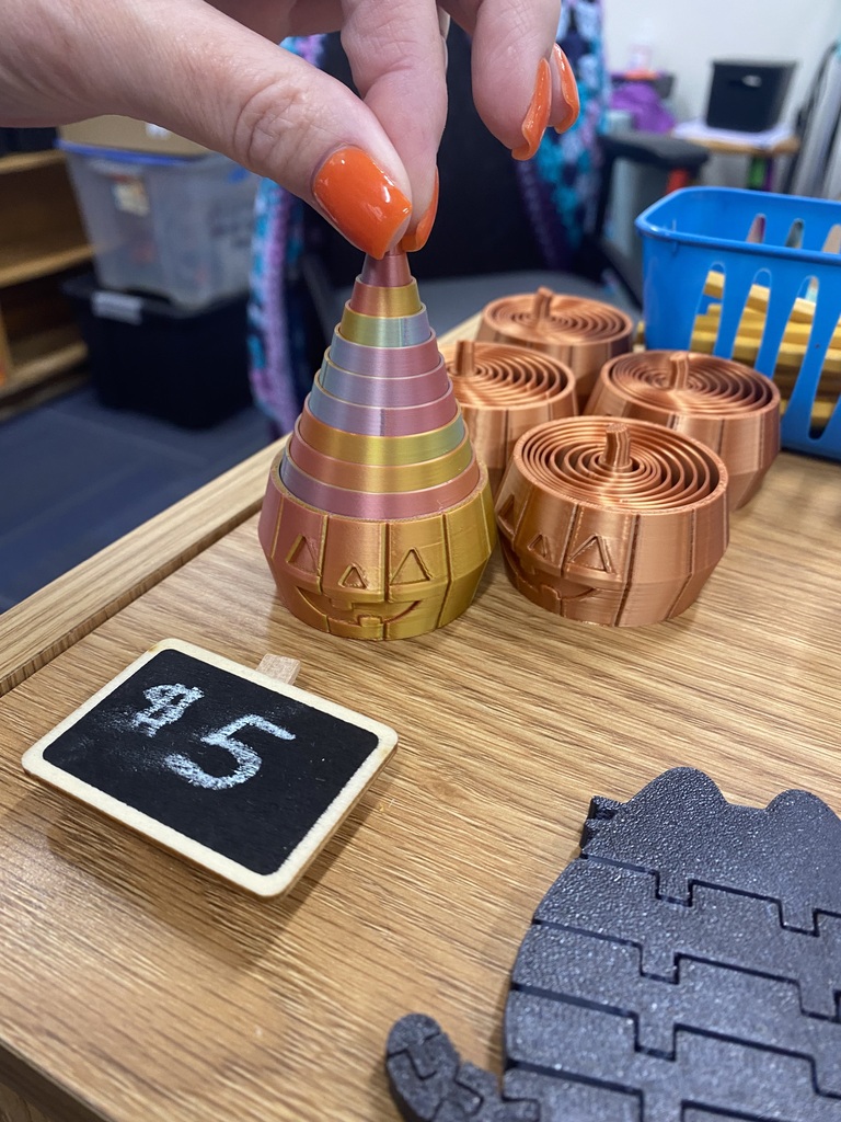 A hand holds a multicolored cone on a table with other items. A chalkboard with the number 5 and a black toy are also on the table.