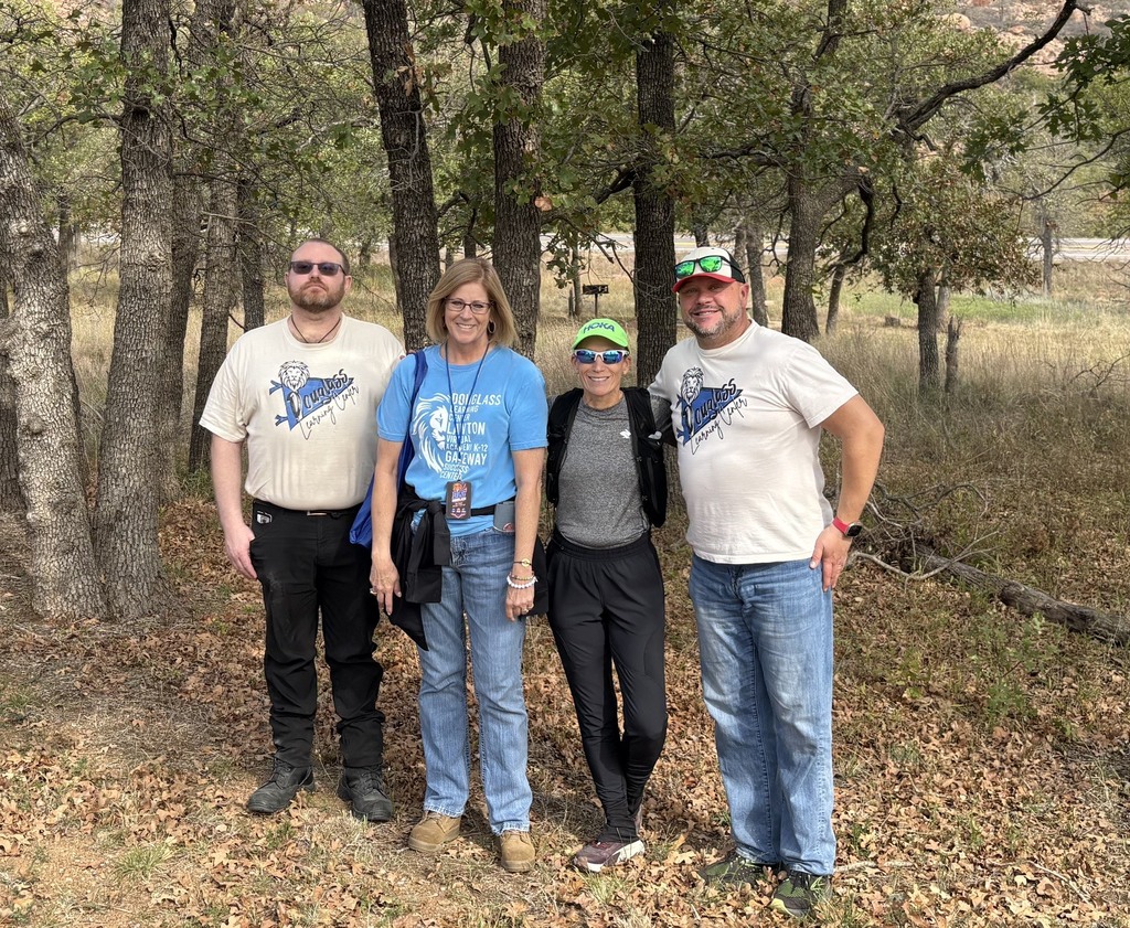 Four people stand in a clearing, surrounded by trees and dry leaves, wearing casual attire.