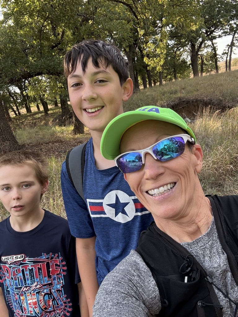 Three people stand in front of a wooded area. A woman wearing sunglasses and a cap smiles.