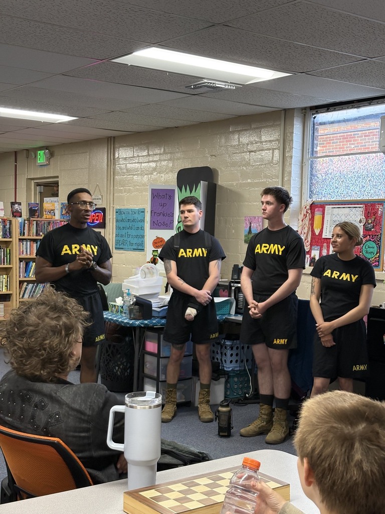 Five people in army uniforms stand in a classroom. One person speaks while others hold objects. People sit at a table. A chess set is on the table.