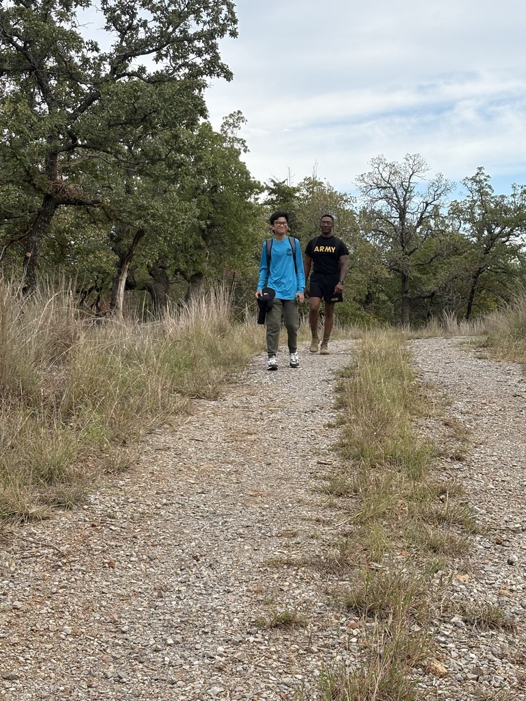 Two students walk on a gravel path surrounded by tall grass and trees.