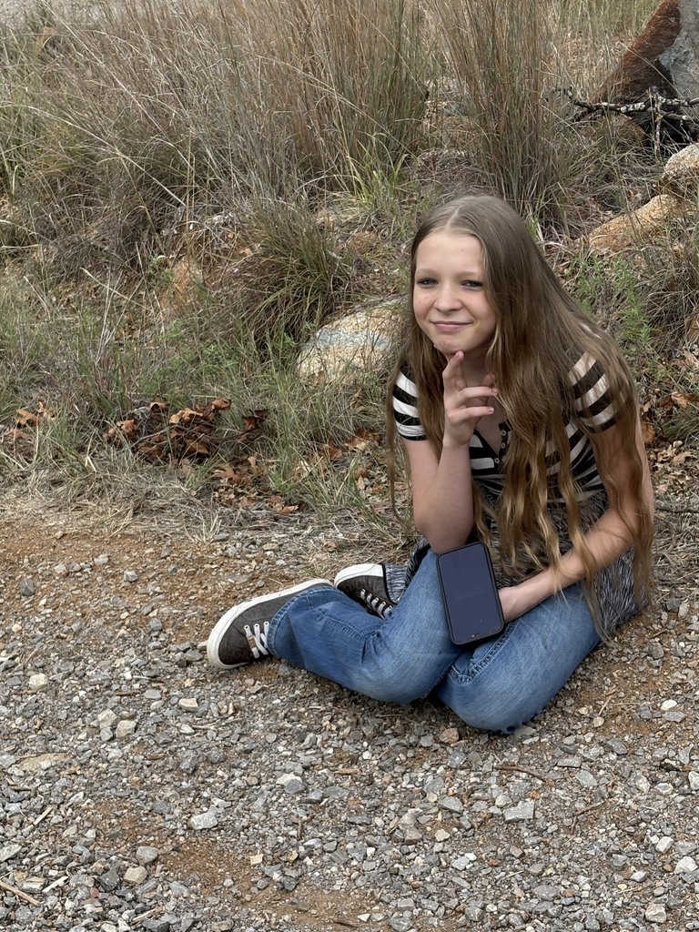 A girl with long hair sits on gravel, holding a phone, and smiling for a photo.