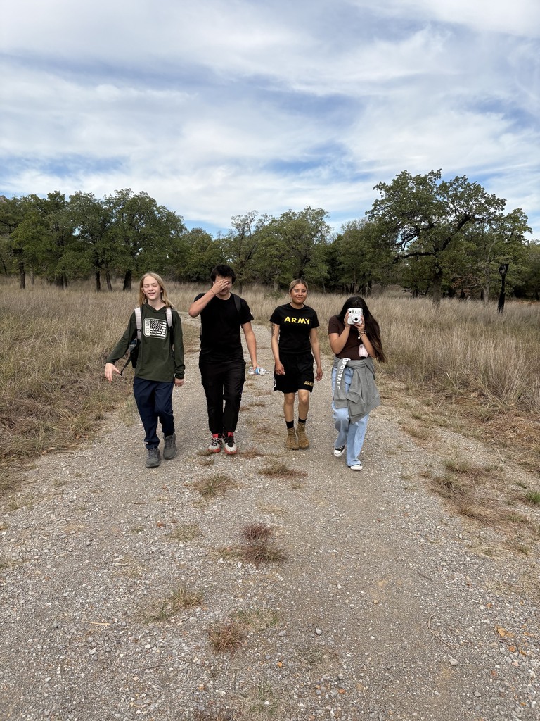 Four individuals walking in a line on a dirt road, surrounded by a grassy field and trees.