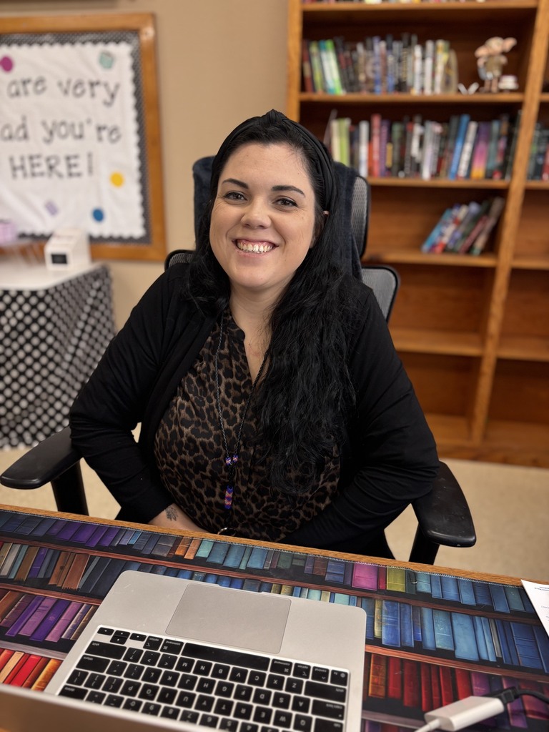 Woman sitting at a desk with a laptop, surrounded by colorful books and a bookshelf.