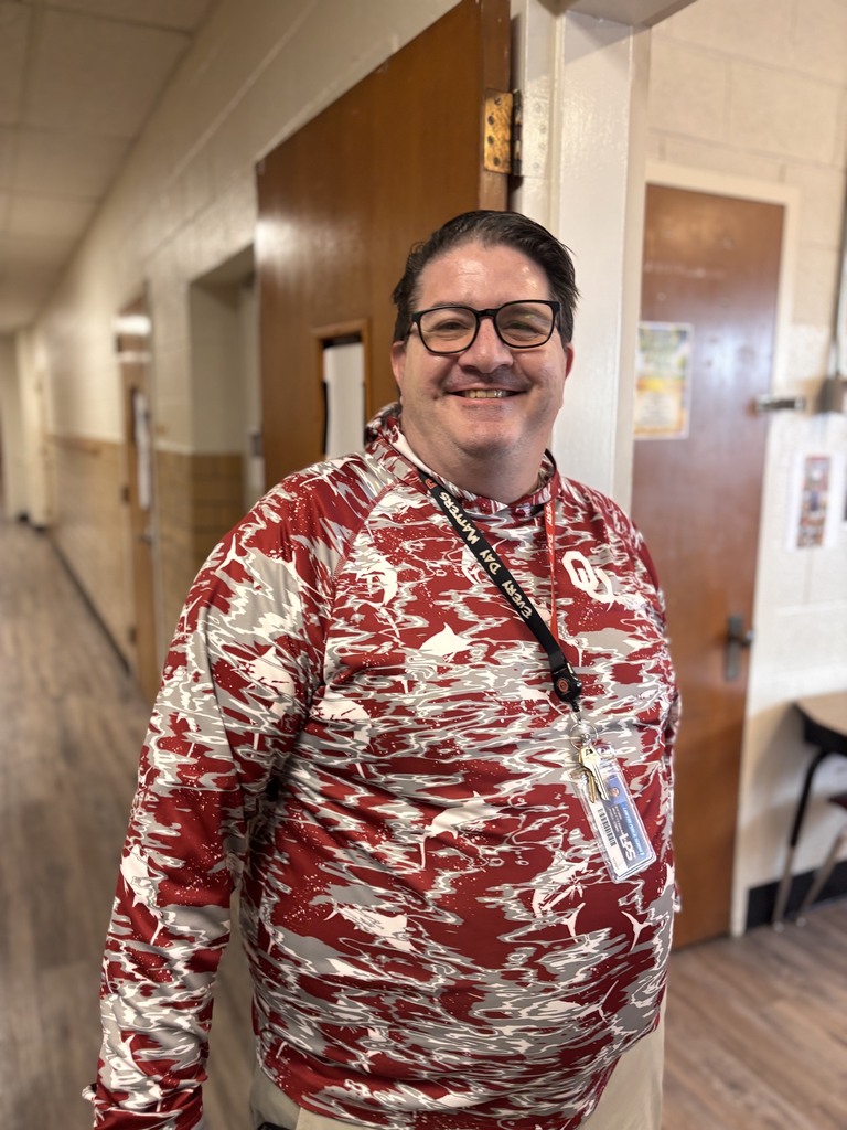Person in red and white patterned shirt and glasses stands in a hallway, smiling at the camera.