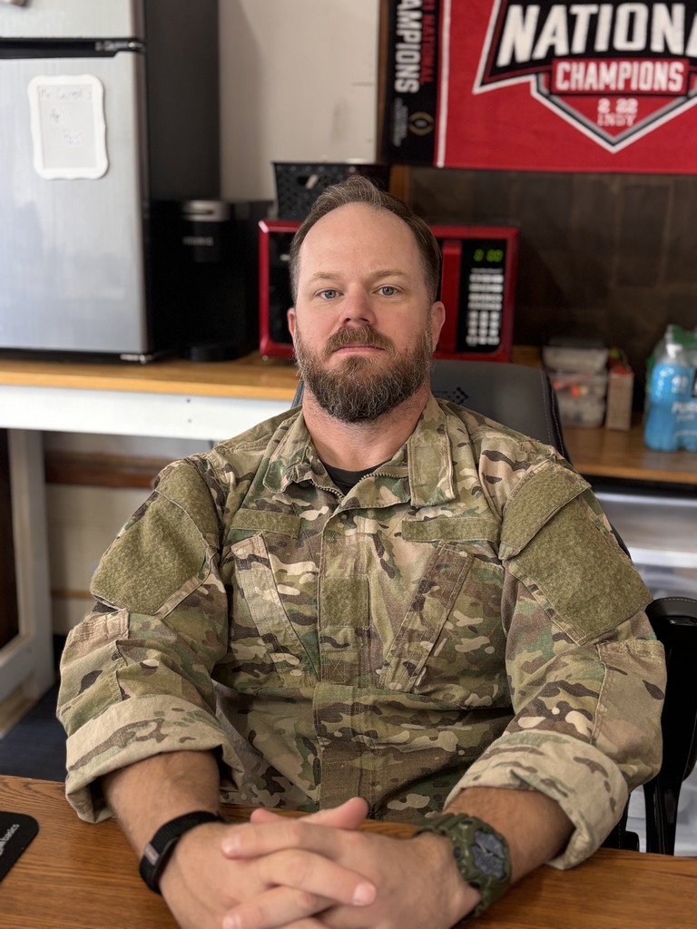A man in a military uniform sits at a desk, his hands clasped in front of him.