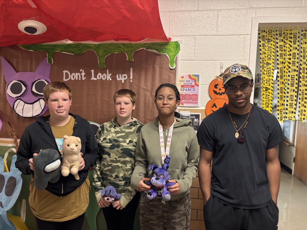 Five young people pose together in front of a Halloween-themed backdrop, each holding a stuffed animal.