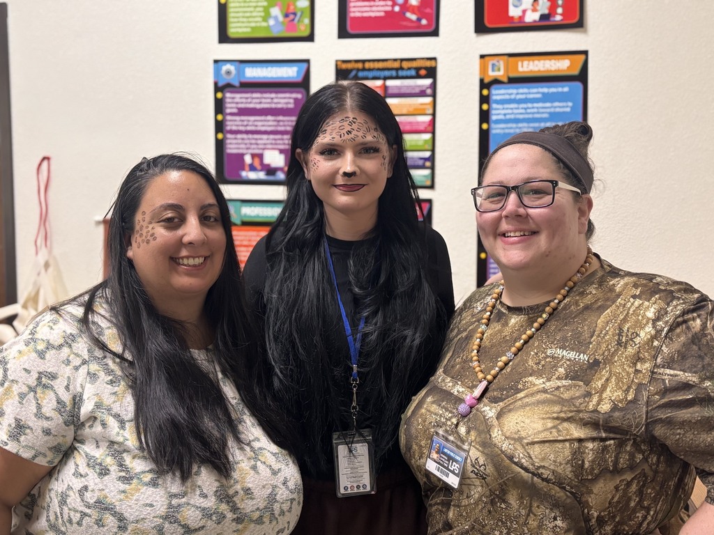 Three adults with face paint stand together in a room with bulletin board behind them.