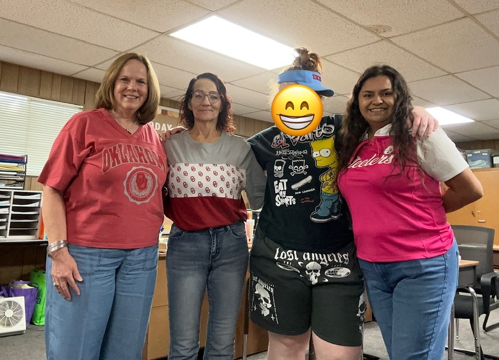 Four women stand together in a room, smiling. The woman on the left wears a red shirt and jeans.