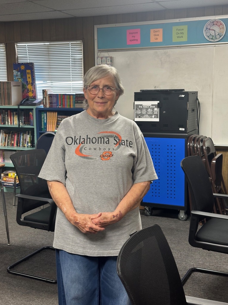 An older woman stands in a classroom, wearing a gray Oklahoma State University T-shirt and jeans. She is surrounded by chairs, a bookshelf, and a printer.