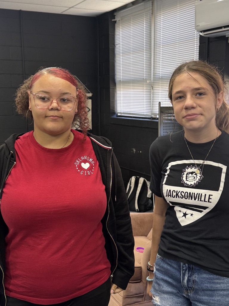 Two women standing, one in a red shirt with a logo, the other in a black shirt. Behind them, a window with blinds, a couch, and a wall.
