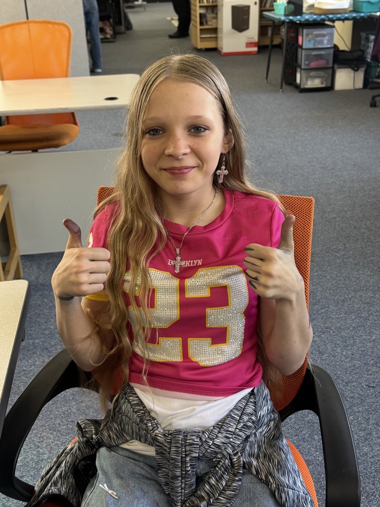 A girl with blonde hair sits in a chair and smiles. She gives a thumbs-up with both hands.