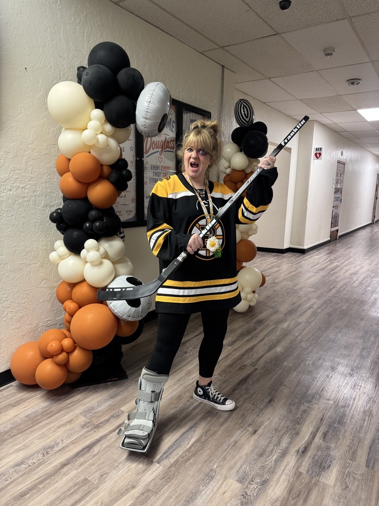Woman with a hockey stick, standing in a hallway with a sports theme. Behind her is a wall with balloons.