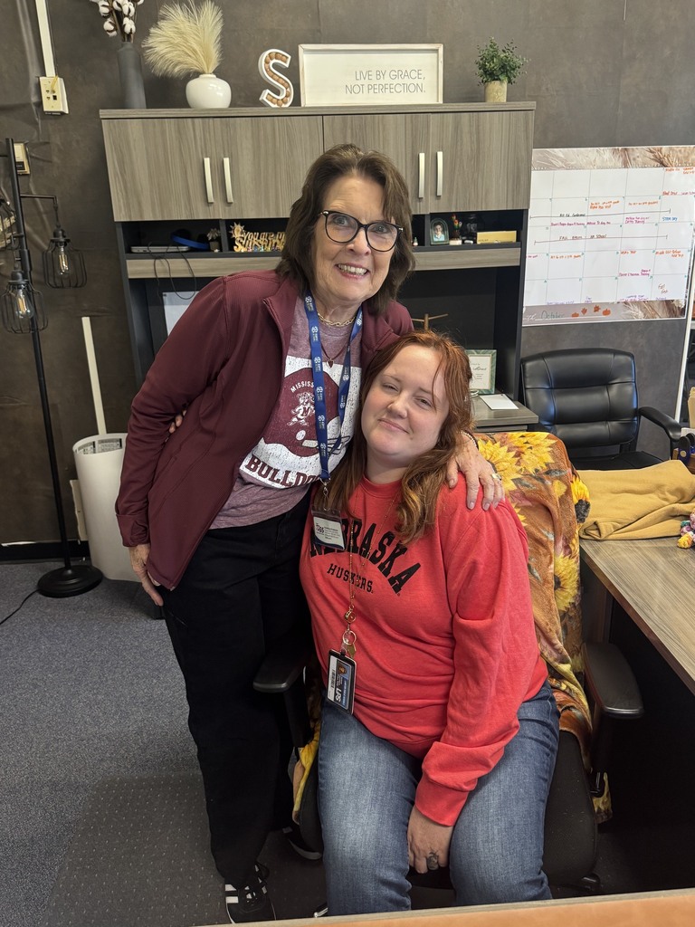 Two women posing in an office; one sitting in a chair, the other standing behind her.