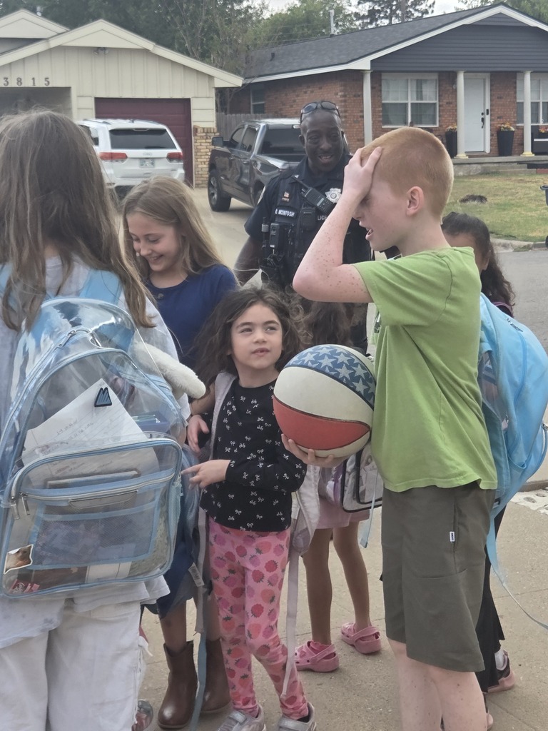 Children stand in a line on a street, with one holding a ball. A person in uniform stands behind them.