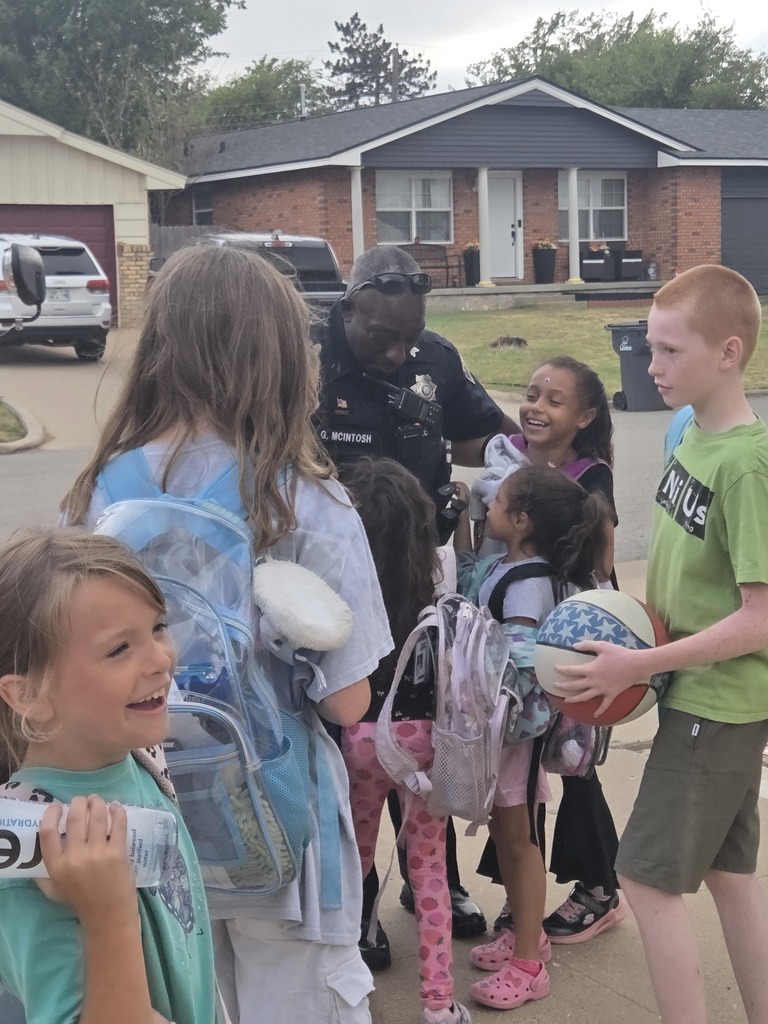 Children gathered, some holding backpacks, a man wearing a police uniform stands with them.