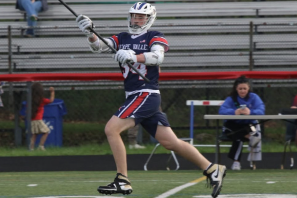 Jaxson Goss runs across the lacrosse field in a Lenape Valley navy, red, and white uniform and helmet, holding his lacrosse stick.