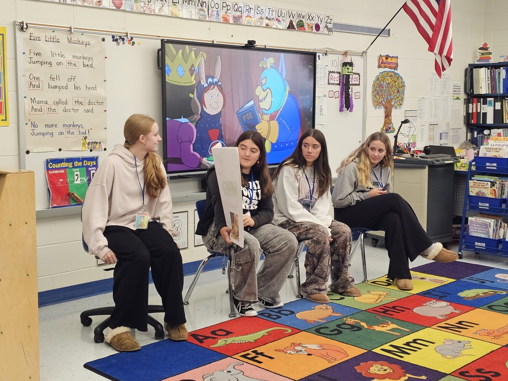 Four Lenape Valley Foundations of Education students sit at the front of a Stanhope Elementary classroom, with one holding up an original children’s book during their storytelling visit.
