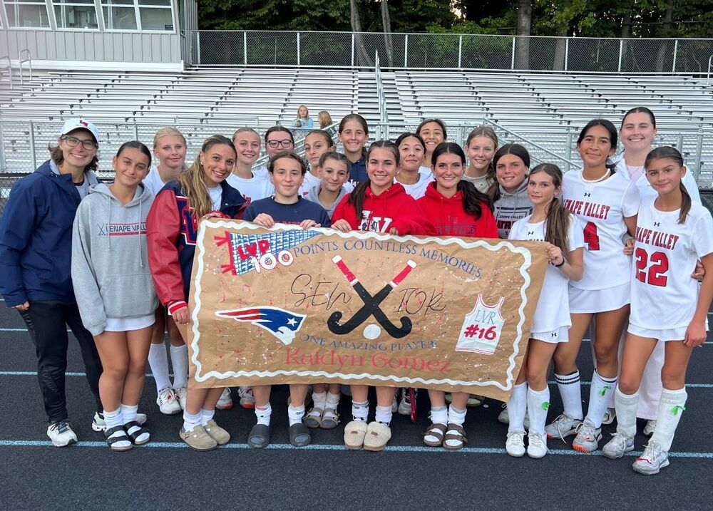 Lenape Valley field hockey player Kaidyn Gomez celebrates reaching 100 career points surrounded by teammates and coaches. The team holds a handmade sign recognizing Gomez and her 100-point milestone.