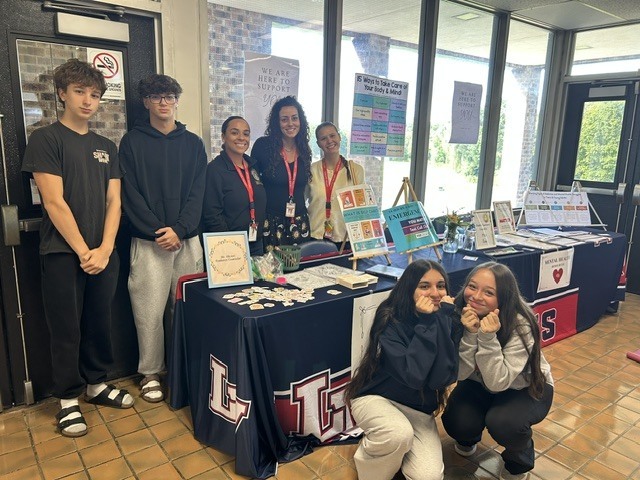 Four students and three school clinicians pose around a Lenape Valley resource table displaying mental health, wellness, and self-care information during a lunchtime student outreach event.