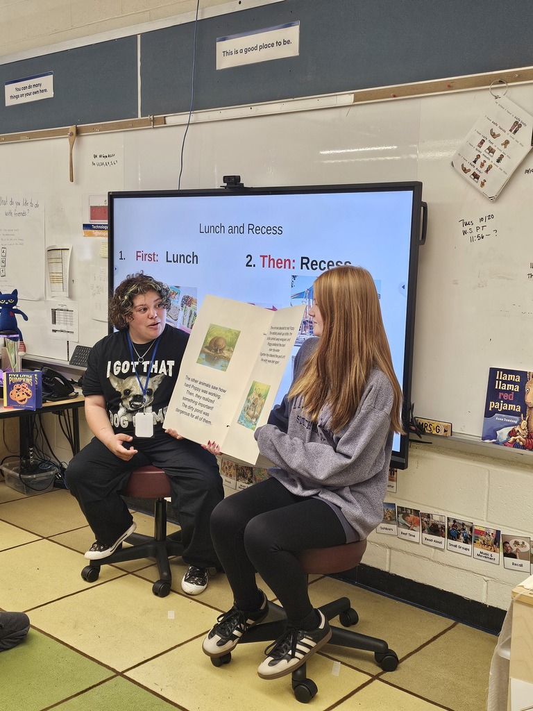 Two Lenape Valley Foundations of Education students sit in a Stanhope Elementary classroom, holding open their original children’s book, “Petite Poppy Platypus,” to show its text and illustrations.
