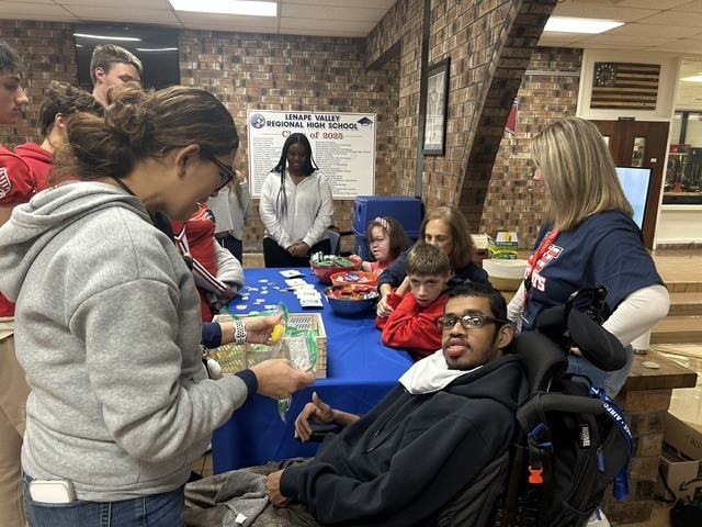 Students and staff gather at the Snacks and Laughs stand as a person holding a basket exchanges items with a student using a wheelchair.