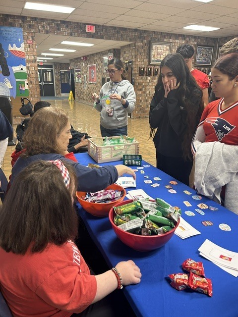 Students and staff interact around the Snacks and Laughs table, displaying colorful stickers, printed cards, and bowls of candy.