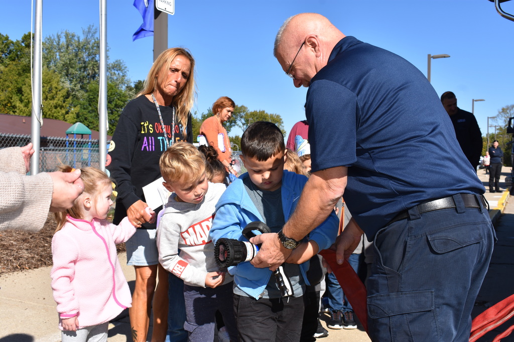 A student holding the hose while others look on and a firefighter helps