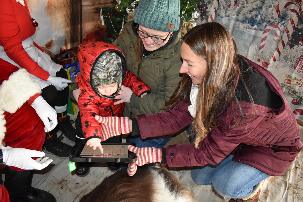 A young boy using his talker to tell Santa what he wants as his teacher and mom assist