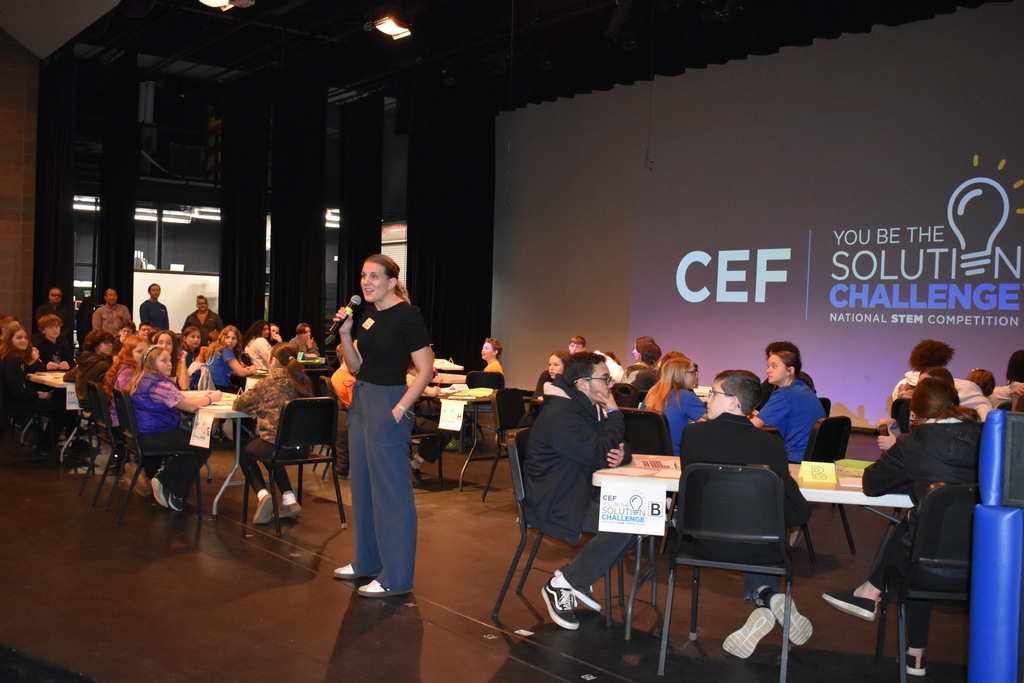 Jackie Murray, presenter, talking on stage with the students placed at their team tables