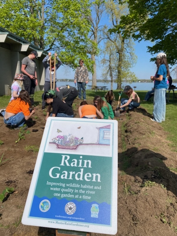 Rain garden sign with students planting rain garden in background.