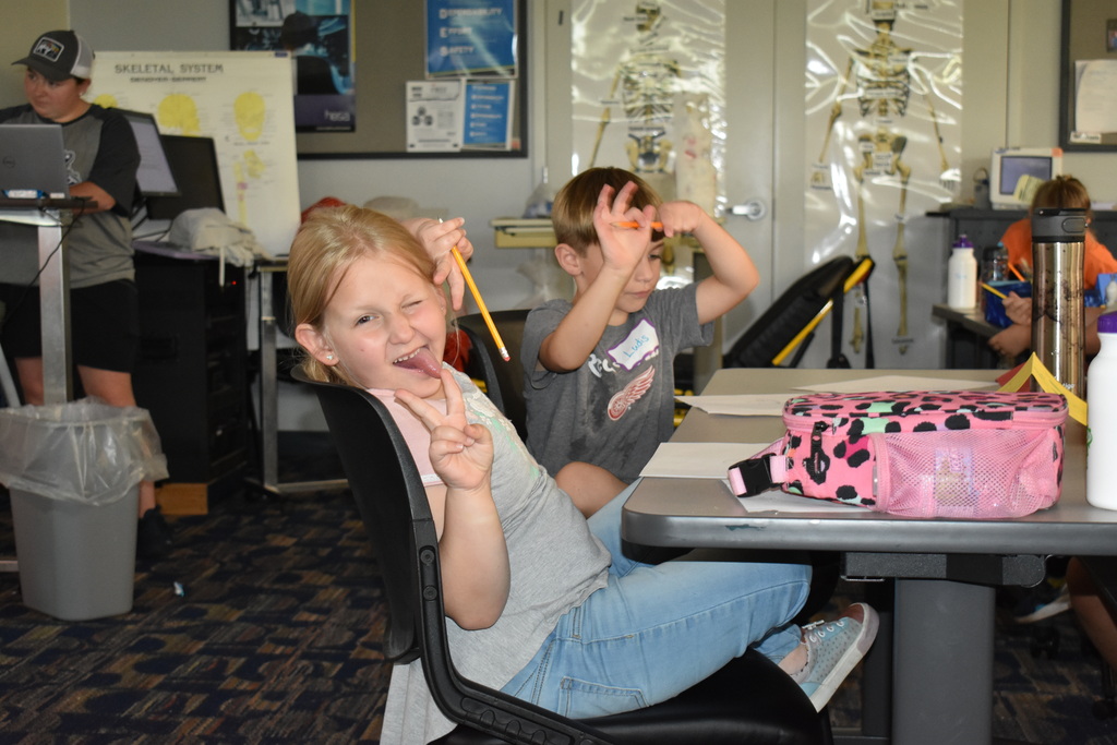 A young girl smiling with her tongue out and giving the peace sign while another student sits behind her and the teacher works at a computer