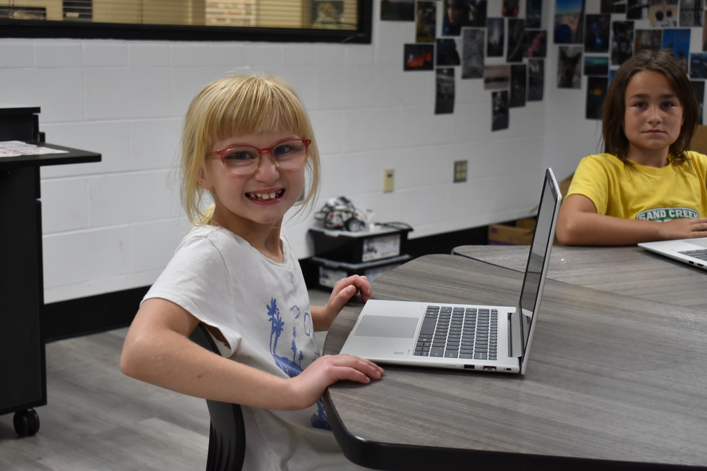 Two students smiling at the camera while seated at a table with their computers