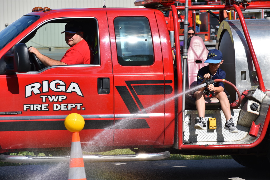 A kid riding on the back of a truck using a fire hose to knock a ball off a cone