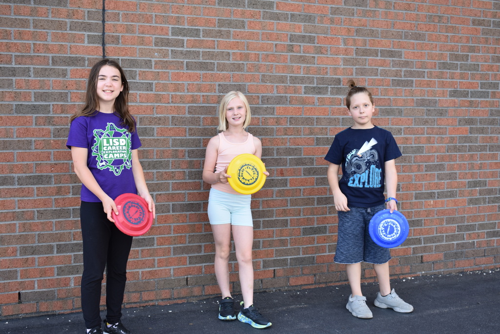 Three kids leaning against a wall and holding frisbees while smiling for the camera