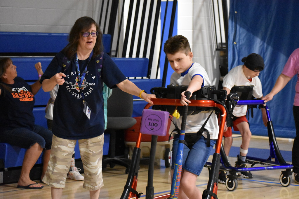 A student swinging the bat at the cube from his assisted walker, as a teacher looks on