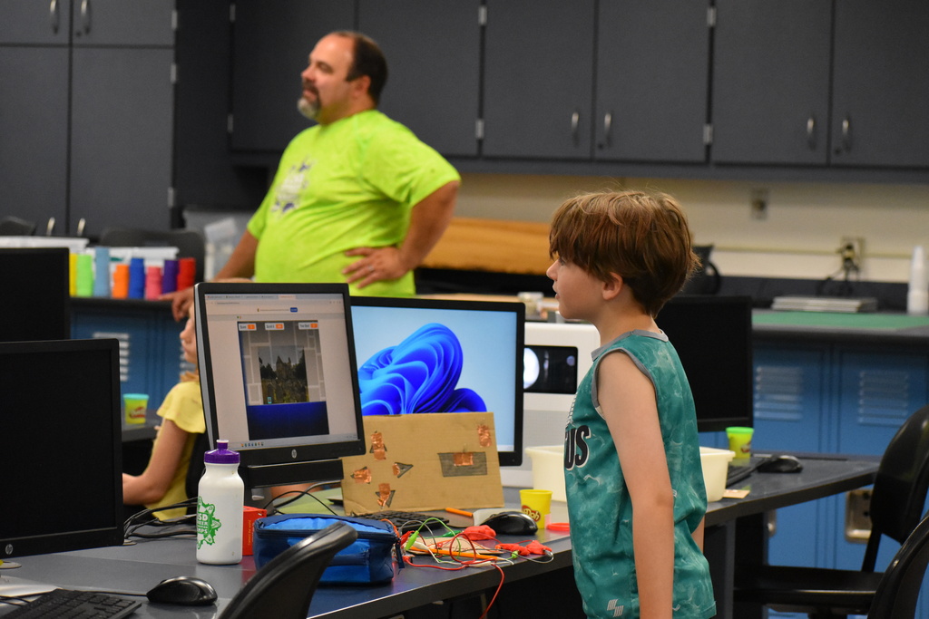 A student working on a computer and a circuit board while a teacher stands in the far background