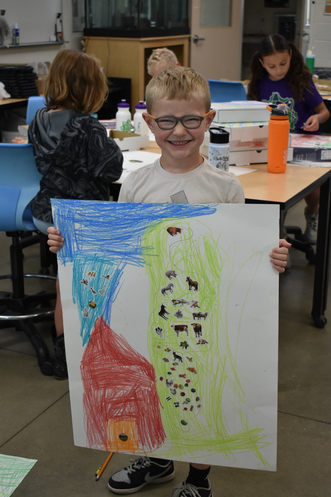 A student smiling at the camera holding his picture of a farm scene that he colored