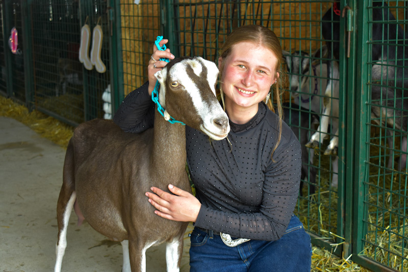 An FFA student smiling at the camera while she is bent down with her arms around her goat inside a barn at the fair