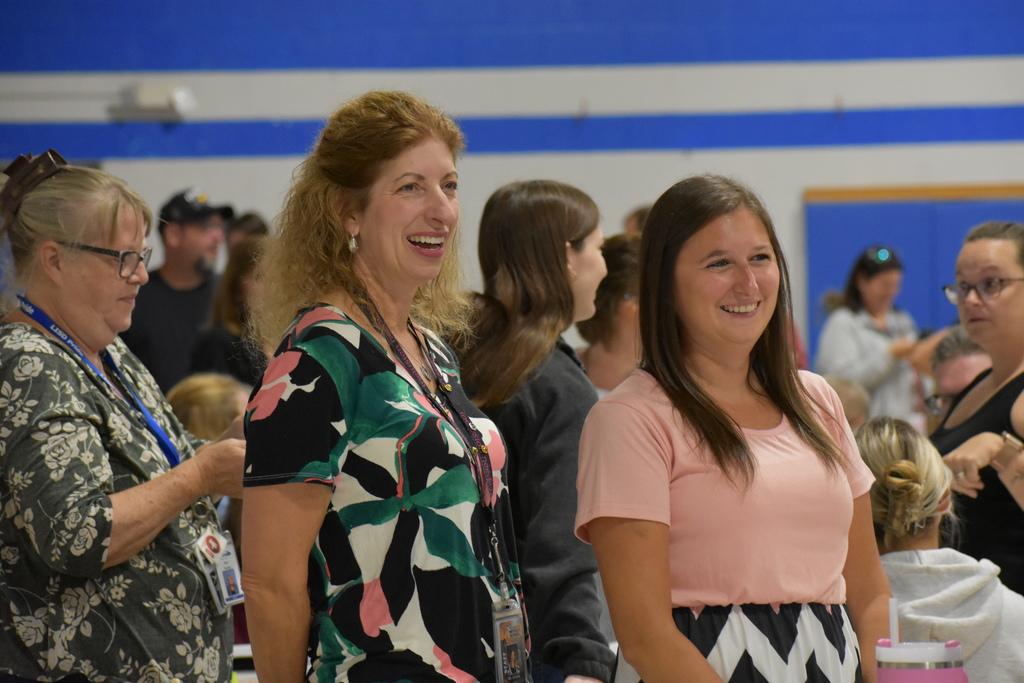 Two staff members smiling and looking at something being presented while standing among their peers in the gymnasium