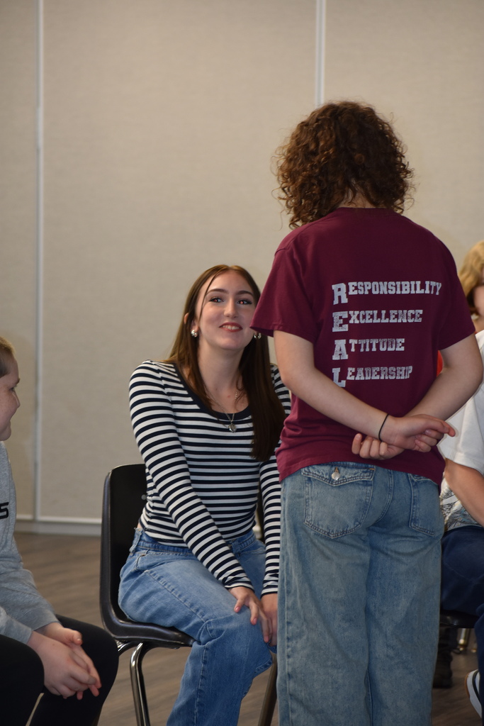 Students seated in a circle for a game and one student standing in front of another asking a question for the game