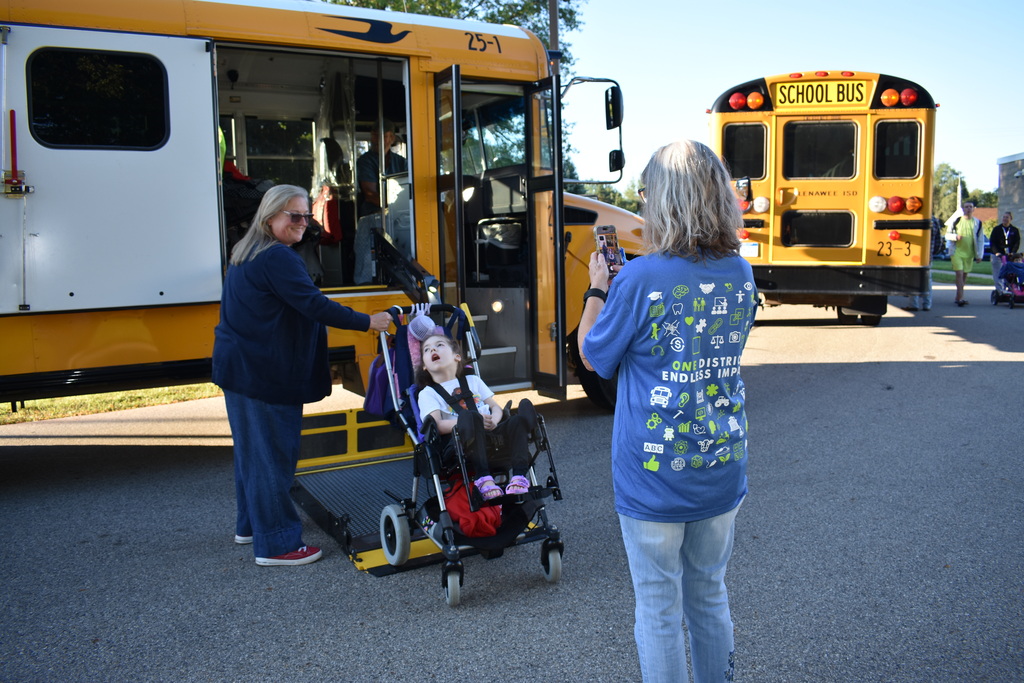 A teacher taking a photo of her student as she comes off the ramp of the bus with the assistance of the bus driver