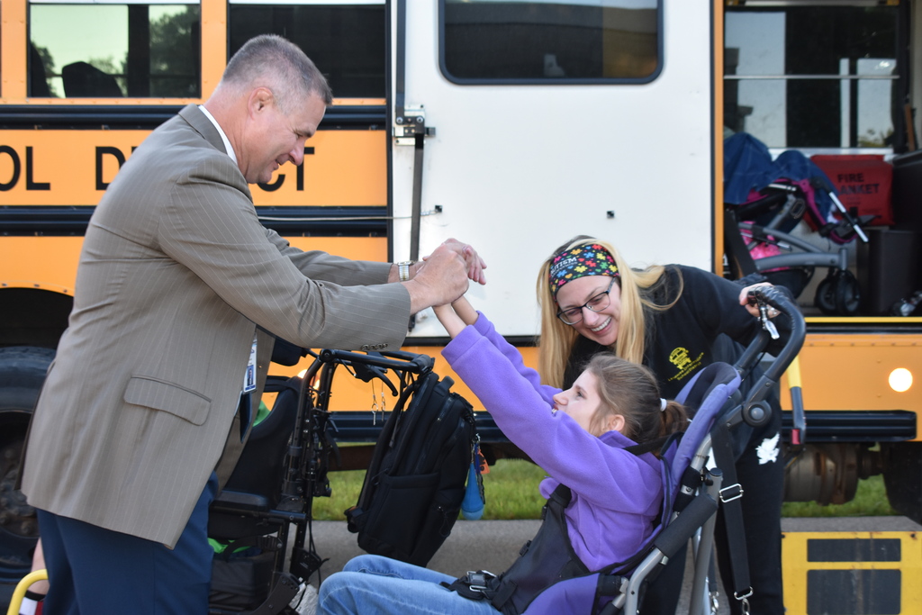 The superintendent holding both hands of a student while her bus driver helps get her settled after exiting the bus ramp