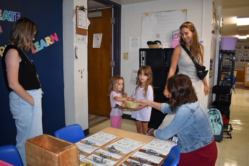 Two young siblings taking paper and a writing tool from a teacher as their parent smiles and looks at the exchange